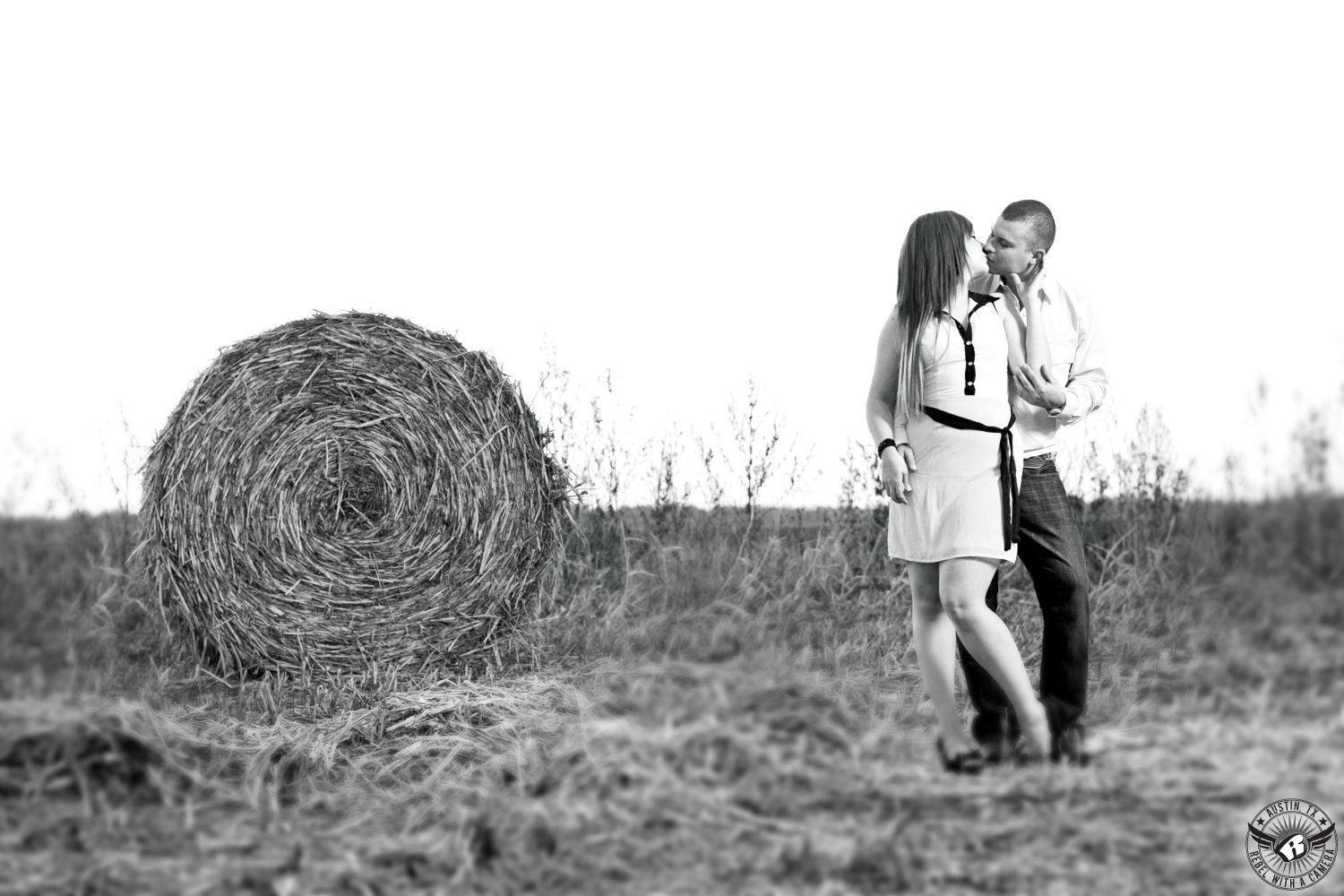 latino girl in light blouse with brown red hair kissing a latino guy wearing a light cowboy shirt  and jeans in a plowed field of corn with a tall round hay bail in the background of this black and white engagement portrait in Granger